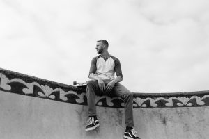 Black and white photo of model Alex Reis sitting on top of skateboard bowl with his skateboard in Santa Cruz