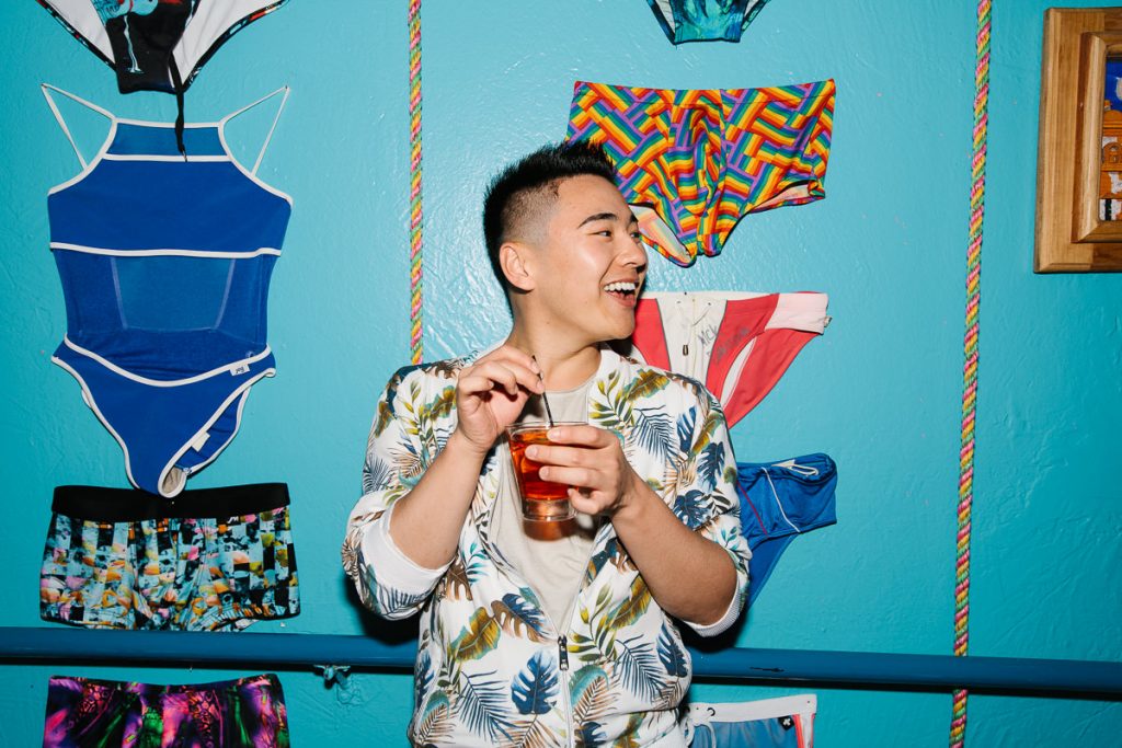 Model holding a drink and laughing in front of a blue wall with swim suits hanging on it for an advertising campaign commissioned by the Campari Group.